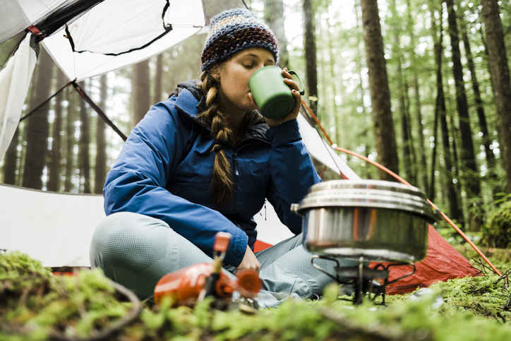 A blonde haired young women sits in front of her camping tent in a lush rainforest with a camp stove and pot steaming in front of her drinks a warm beverage from a mug.