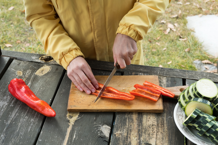A woman in a bright yellow jacket slices zucchini and bell peppers to cook them on the grill, on an autumn sunny day. Vacation or vacation in a country house, on a picnic or camping. You cook on coals, on fire, by yourself.